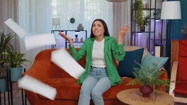 Stressed girl at home room looking at unpaid bank debt bills, doing paperwork, planning budget, calculate finances mortgage payments. Displeased young woman throws paper bills sitting on orange couch