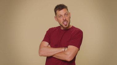 Cheerful funny bully young man in red t-shirt. Adult guy showing tongue making faces at camera, fooling around, joking, aping with silly face, teasing isolated alone on beige studio background indoors