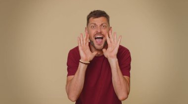 Expressive bearded handsome young man holding hands near mouth loudly screaming, shouting, yelling, announces discounts sale Hurry up, welcomes someone hello. Adult stylish guy on beige background