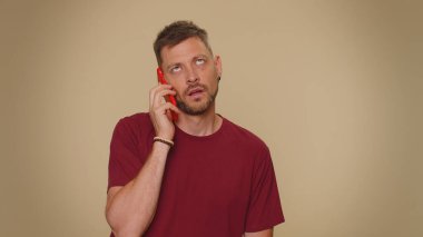 Tired young man in red t-shirt making online conversation speaks on smartphone. Boring call. Blah expression. Nonsense talking. Adult stylish male guy isolated alone on beige studio wall background
