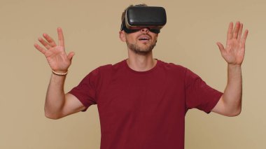 Bearded handsome young man using headset helmet app to play simulation drawing game. Watching virtual reality 3D 360 video. Adult male guy in VR goggles isolated on beige studio background indoors