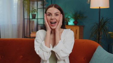 Oh my God, Wow. Lovely adult girl surprised looking at camera with big eyes, shocked by sudden victory, good win news, celebrating at home. Excited amazed young woman indoors in living room on couch