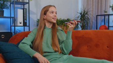 Portrait of caucasian girl making phone conversation with friends sitting call on couch at home in room. Happy excited young child kid enjoying mobile loudspeaker talking indoors at home apartment