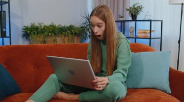 Overjoyed student girl child kid at home couch with laptop scream in delight raise hands in triumph winner gesture celebrate success win money in lottery. Excited young woman get online good news