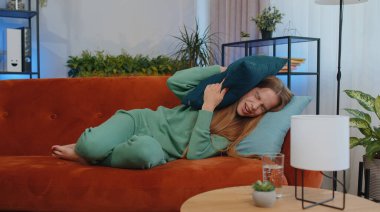 Repair work at neighbours concept. Irritated girl sit on couch cover ears with pillows annoyed by noisy neighbors suffer from headache wishes silence. Thin walls at home flat without sound insulation