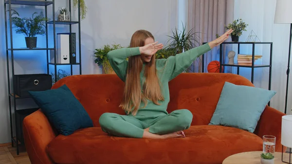 Trendy cheerful positive teen girl having fun dancing, moving to rhythm, dabbing raising hands, making dub dance gesture. Young caucasian child kid at modern home apartment living room on sofa couch