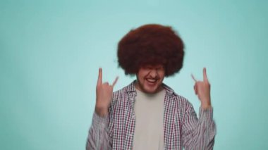 Rock n roll. Overjoyed delighted man 30 years old in shirt showing gesture by hands, cool sign, shouting yeah with crazy expression, dancing, emotionally rejoicing in success win alone on blue wall