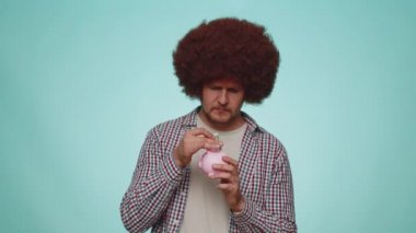 Poor man upset over insufficient amount of money, holding piggybank and one dollar banknote. Financial crisis. Bankruptcy. Poverty and destitution. Young guy isolated on blue studio wall background