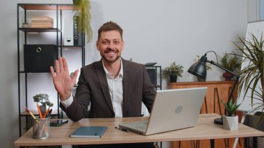 Businessman in suit working on laptop computer smiling friendly at camera and waving hands gesturing hello, hi, greeting or goodbye, welcoming with hospitable expression at office workplace desk
