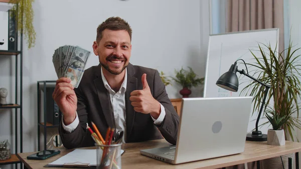 Happy businessman in suit working on laptop computer sincerely rejoicing win, receiving money dollar cash banknotes, success lottery luck at office workplace desk. Professional manager freelancer man