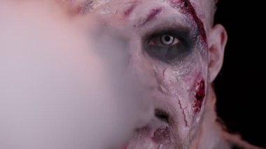 Cadılar Bayramı zombisi makyajlı korkutucu bir adam. Korkutmaya çalışıyor, burnundan ve ağzından duman üflüyor. Korku teması. Stüdyonun siyah arka planında izole edilmiş uğursuz ölümsüz adam. Voodoo ritüelleri
