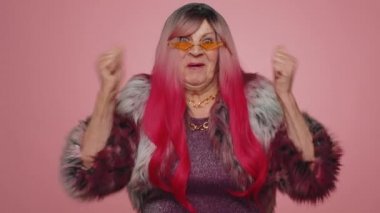 Palto giymiş, bağıran, el kol hareketi yapan mutlu yaşlı bir kadın. Başarımı, kazanmamı, doğum günümü, piyango vuruşumu kutladım. Pembe stüdyo arka planında yaşlı bir büyükanne.