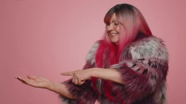 Güzel, şık, yaşlı bir kadın boş bir yeri işaret ediyor. Ticari metinler için reklam alanı, ürün tanıtımı için fotokopi alanı. Yaşlı büyük anne. Pembe arka planda kapalı stüdyo