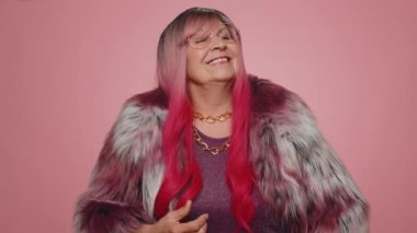 Gülümseyen, mutlu, paltolu, göz kırpan yaşlı kadın dişlek gülümsemesiyle kameraya bakıyor, göz kırpıyor ve flört ediyor, iyimserliğini ifade ediyor. Yaşlı bir büyükanne pembe duvar arkasında tek başına.