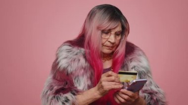 Para transferi, online alışveriş, ödeme yaparken kredi kartı ve akıllı telefon kullanan saygıdeğer yaşlı kadın müşteriler. Finans ve internet. Yaşlı büyükanne pembe arka planda izole edilmiş.