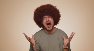 Man with lush brown hairstyle screams from stress tension problems feels horror hopelessness fear surprise shock expresses gestures rage. Young guy boy isolated alone on beige studio wall background