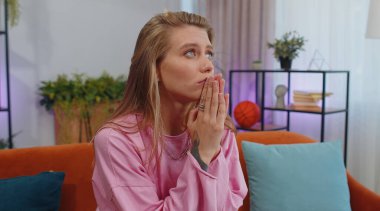 Please, God help. Girl praying sincerely with folded arms, looking upward and making wish, asking with hopeful imploring expression, begging apology. Young woman sitting on couch at home living room