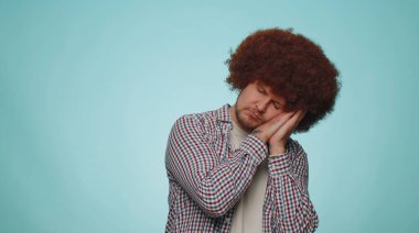 Tired handsome bearded man with lush hair coiffure yawning, sleepy inattentive feeling somnolent lazy bored gaping suffering from lack of sleep. Young guy boy isolated alone on blue studio background