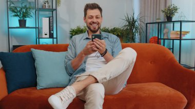 Portrait of man use mobile smartphone typing browsing say Wow yes found out great big win good news celebrate lottery jackpot doing winner gesture. Young guy at home in living room sitting on sofa