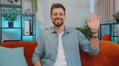 Portrait of caucasian adult man smiling friendly at camera and waving hands gesturing hello, hi, greeting or goodbye, welcoming with hospitable expression. Young guy indoor at home in room on couch