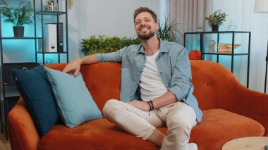Portrait of happy calm adult man in shirt smiling friendly, glad expression looking away dreaming resting, relaxation feel satisfied concept good news. Young caucasian guy sitting on sofa at home room