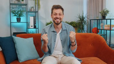 Happy caucasian man shouting, celebrating success, winning, goal achievement, good news, raising fists in gesture I did it, lottery luck at home apartment. Young guy indoors in living room on couch