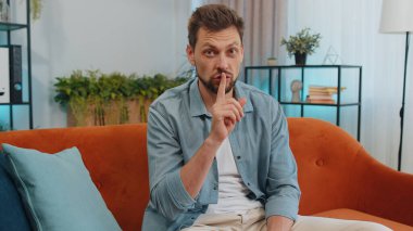 Sessiz olun lütfen. Yetişkin beyaz bir adam işaret parmağını dudaklarına bastırır sessiz ol işareti yapar dedikodu sırrı söylemez. Evdeki genç adam oturma odasında turuncu koltukta oturuyor.
