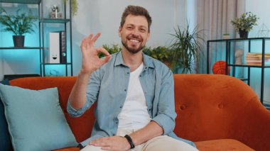- Tamam. Mutlu, neşeli, yakışıklı bir adam kameraya bakıp, olumlu bir hareket gösteriyor, işaret gibi, oturma odasında turuncu koltukta oturan güzel bir şeyi onaylıyor. Modern ev dairesindeki genç adam.