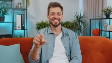 Hey, sen! Yetişkin bir adamın portresi heyecanlı bir şekilde gülümsüyor ve kamerayı işaret ediyor, şanslı kazananı seçiyor, sizi davet ediyor, onaylıyor. Modern ev dairesindeki kanepede oturan genç adam.