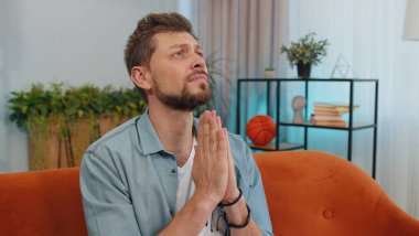 Please, God help. Man praying sincerely with folded arms, looking upward and making wish, asking with hopeful imploring expression, begging apology. Young guy sitting on couch at home living room