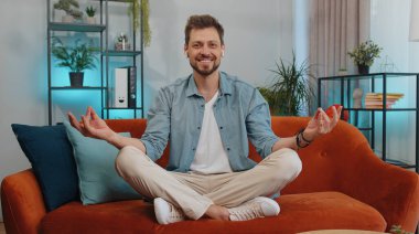 Sakin ol, rahatla, iç denge. Yetişkin adam derin nefes alıyor Mudra jesti ile, gözleri kapalı meditasyon yapıyor konsantre düşüncelerle, huzurlu zihinle. Genç adam oturma odasında kanepede oturuyor.