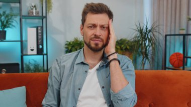 Kafkasyalı yetişkin bir adamın baş ağrısı, tansiyon, migren, stres, ekşitme, yüksek tansiyon gibi hastalıkları tedavi etmek için tapınakları ovmasından memnun değil. Evdeki kanepede oturan genç adam.