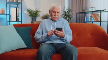 Kanepede oturan yaşlı büyükbaba modern ev dairesinde cep telefonuyla gülümsüyor. Çevrimiçi akıllı telefon sosyal medya uygulamaları üzerinden mesajlaşan yaşlı bir adam, rahatlama filmi izliyor