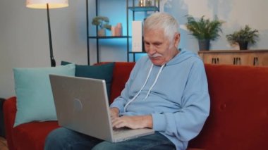 Overjoyed senior old grandfather on home couch with laptop scream in delight raise hands in triumph winner celebrate success win money in lottery. Excited elderly retired man get online good news