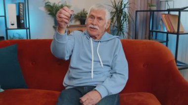 Senior old grandfather real estate agent lifting hand showing the keys of new home house apartment, buying or renting property, mortgage loan at modern home apartment indoors. Man in room sits on sofa