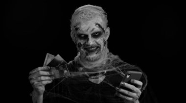 Korkunç korkunç Cadılar Bayramı zombi makyajlı kötü adam kredi kartı ve akıllı telefon kullanarak online alışveriş yapıyor. Yaralı, yaralı yüzlü, siyah odalı ölü bir adam.