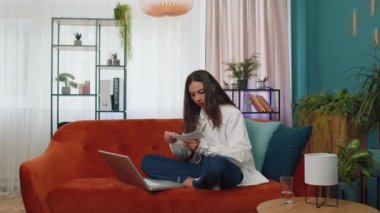 Stressed girl at home room looking at unpaid bank debt bills, doing paperwork, planning budget, calculate finances mortgage payments. Displeased young woman throws paper bills sitting on orange couch