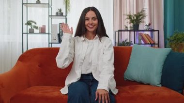 Portrait of caucasian adult girl smiling friendly at camera and waving hands gesturing hello, hi, greeting or goodbye, welcoming with hospitable expression. Young woman indoor at home in room on couch