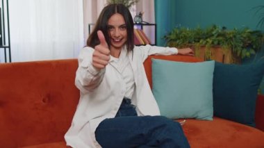 Like. Happy lovely adult girl looking approvingly at camera showing thumbs up, like sign positive something, good news, positive feedback. Young woman sitting on orange couch at home living room
