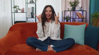 Overjoyed delighted caucasian adult girl showing rock n roll gesture by hands, cool sign, shouting yeah with crazy expression, dancing, emotionally rejoicing in success. Young woman at home apartment