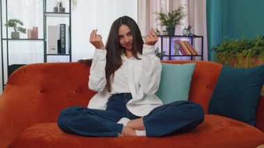 Portrait of caucasian cheerful rich girl showing wasting or throwing money around hand gesture, more tips earnings, big profit, win lottery, body language, share. Young woman at home in room on couch