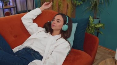 Young woman in wireless headphones relaxing lying on sofa at home choosing listening favorite energetic disco rock n roll dancing music in smartphone. People weekend activities. Happy overjoyed girl