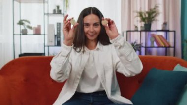 Girl freelancer holding two gold coins at home, successful developer programmer. Woman stock trader earning bitcoins after online monitoring trading operations. Increasing wealth, financial prosperity