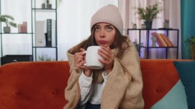 Sick girl wear hat wrapped in plaid sit alone shivering from cold on sofa drinking hot tea in unheated apartment without heating due debt. Unhealthy young woman feeling discomfort try to warming up