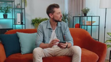 Kanepede oturan yetişkin bir adamın portresi modern ev dairesinde cep telefonu kullanıyor. Çevrimiçi akıllı telefon sosyal medya uygulamaları üzerinden mesajlaşan genç adam, rahatlama filmi izliyor