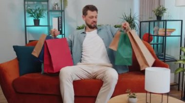 Portrait of caucasian adult man happy shopaholic consumer came back home after shopping sale with bags. Young guy satisfied received parcels from online order at modern home apartment living room