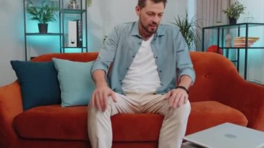 Handsome adult man freelancer enters the home living room and sits down on orange couch, opens laptop. Young guy works on notebook, sends messages, makes online purchases, watching movies, working