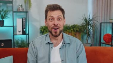 Oh my God, Wow. Handsome adult man surprised looking at camera with big eyes, shocked by sudden victory, good win news, celebrating at home. Excited amazed young guy indoors in living room on couch