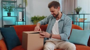Portrait of happy adult man unpacking delivery parcel sitting at home. Smiling satisfied young guy shopper, online shop customer opening cardboard box receiving purchase gift by fast postal shipping
