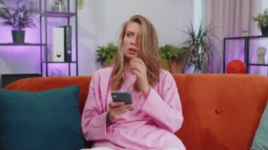 Portrait of adult girl sitting on sofa uses mobile phone smile at modern home apartment. Young woman texting share messages content on smartphone social media applications online, watching relax movie